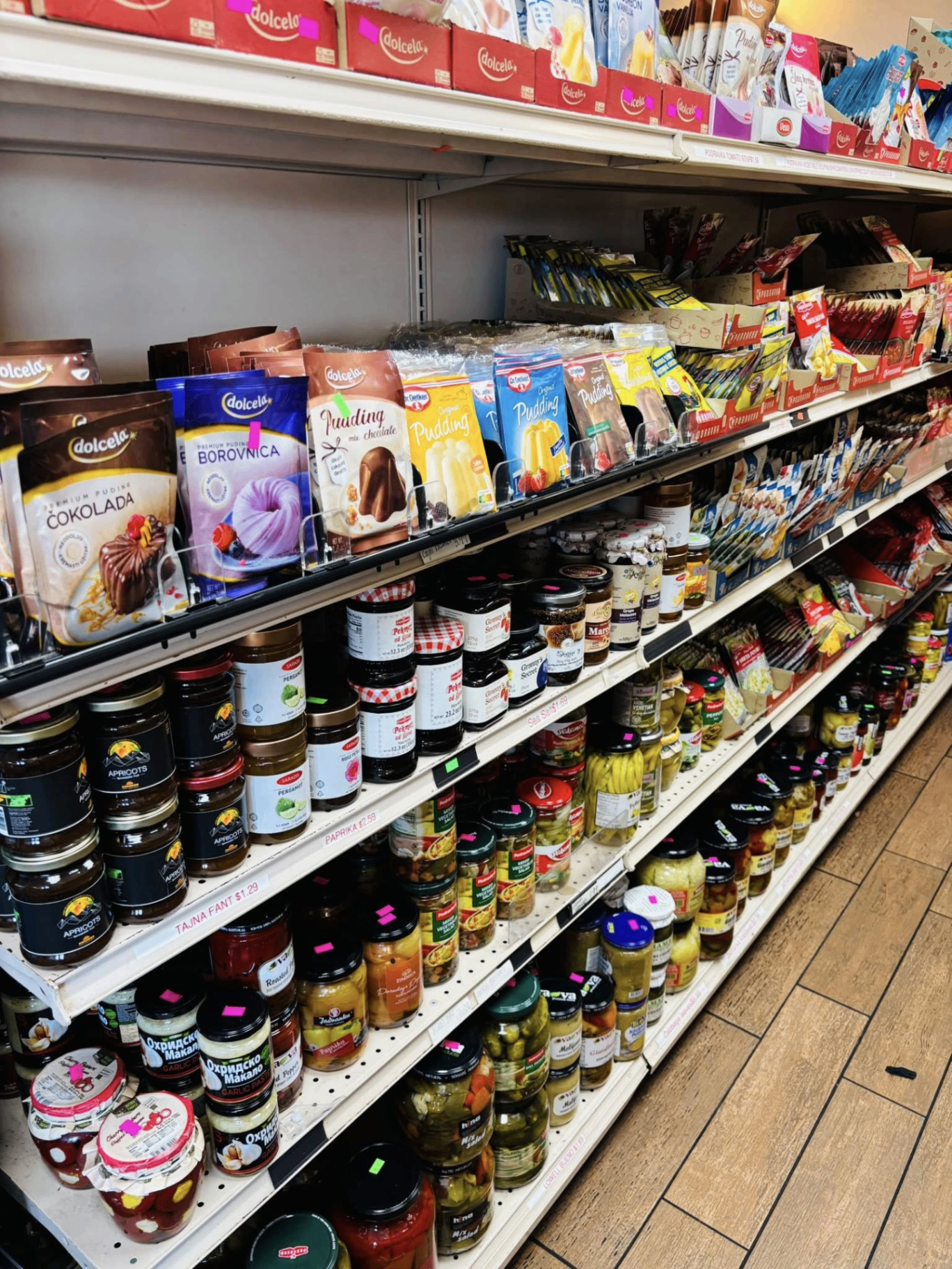 Market shelves stacked with imported European and Bosnian groceries — ajvar, pudding, pickled vegetables and sweets